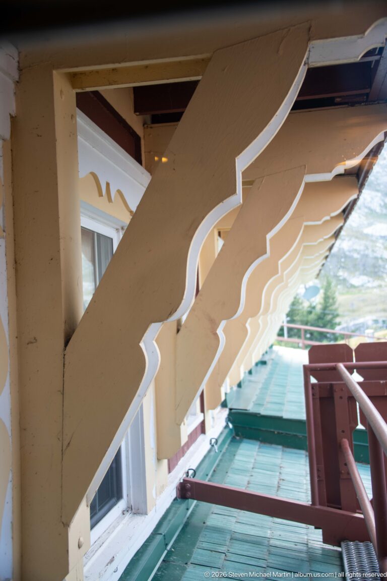 Row of large curved wooden balcony brackets with light trim above green metal roofing and red-brown railings.