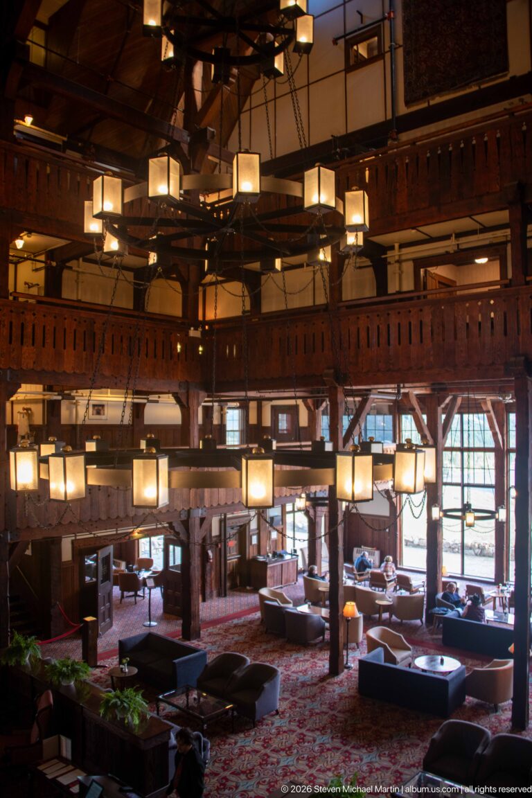 Upper-level view across the timber hotel lobby with hanging lights, sofas, tables, balconies, and bright windows.