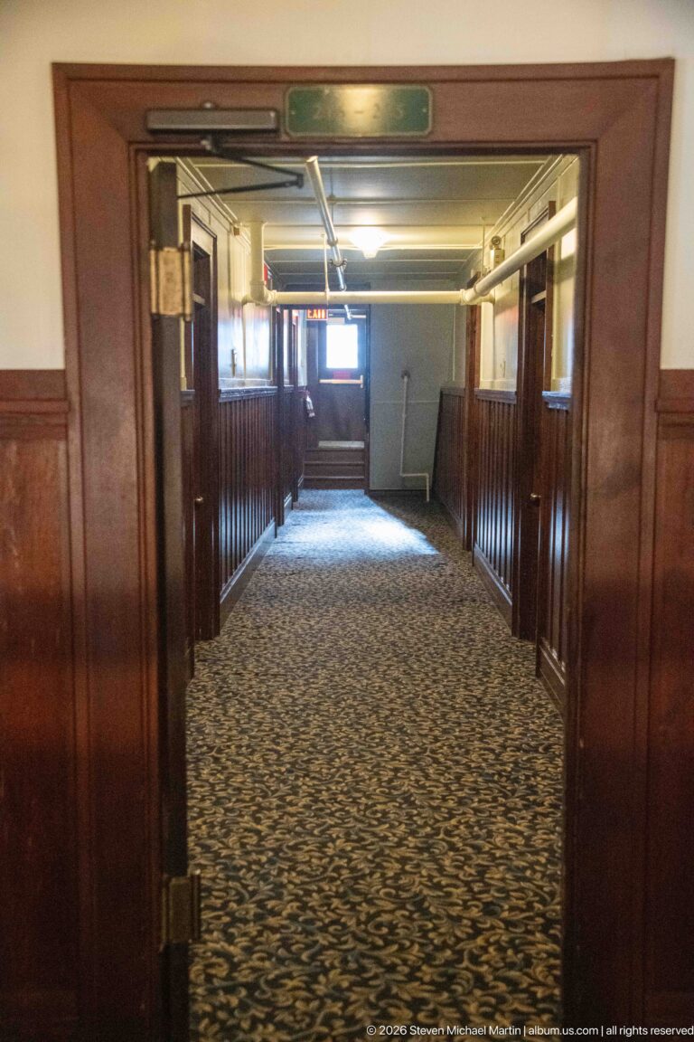 Long hotel corridor with patterned carpet, dark wood wainscoting, exposed ceiling pipes, and a bright window at the far end.