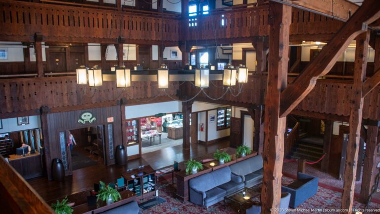 Elevated view of timber posts, hanging lights, sofas, plants, service openings, and staircase in the hotel lobby.
