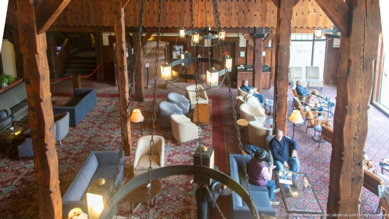 Guests sit among sofas and armchairs between massive timber columns, viewed from above beside a large chandelier.