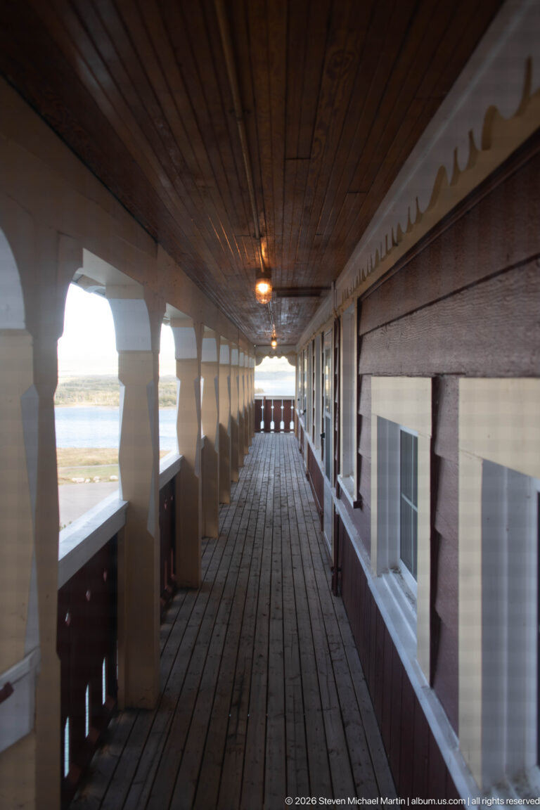 Long covered wooden hotel gallery with plank floor and ceiling, repeating posts, openings, and guest-room windows.