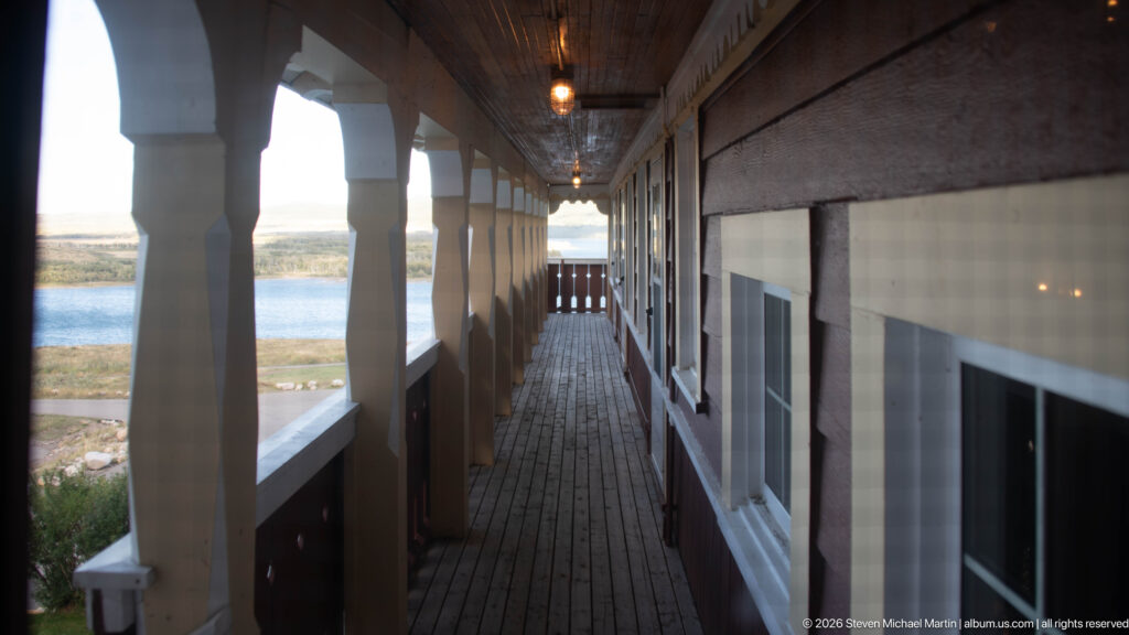 Long covered wooden hotel gallery with repeating posts, guest-room windows, plank floor, and glimpses of lake and landscape.