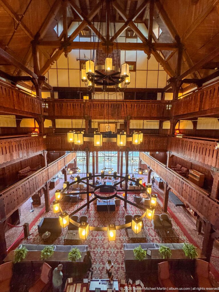 Elevated symmetrical view of the timber hotel lobby with stacked balconies, roof trusses, chandeliers, patterned carpet, and lounge seating.