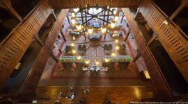 Top-down view through a circular chandelier onto sofas, tables, plants, patterned rugs, and the timber lobby floor.