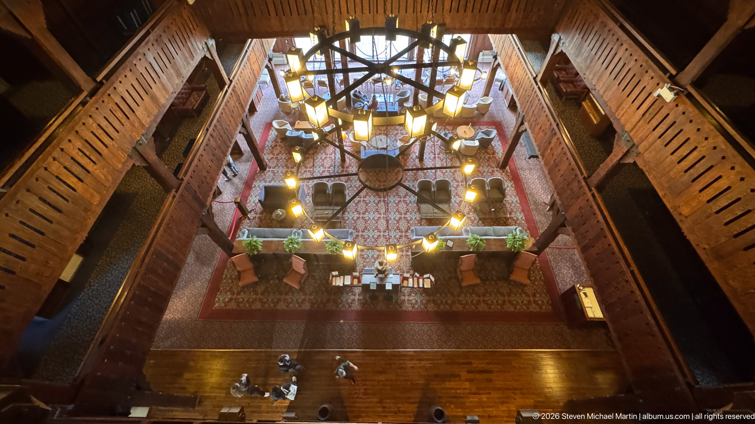 Top-down view through a circular chandelier onto sofas, tables, plants, patterned rugs, and the timber lobby floor.